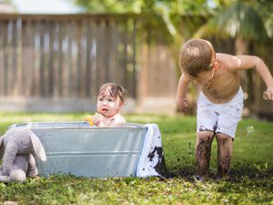 Baby taking a bath in metal tub with brother playing in the mud next to him